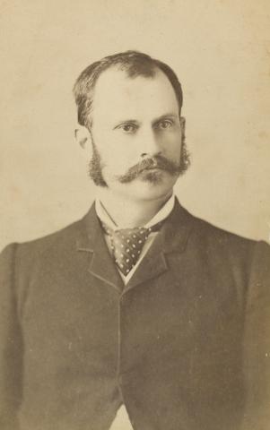 Bust length photo of a man in a black suit. His hair is brown with muttonchop sideburns and a large moustache.