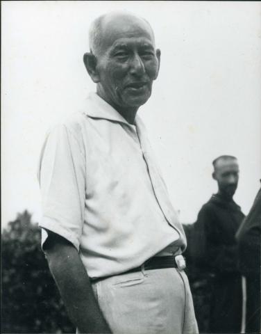 ¾ length photo of a balding man wearing a white short-sleeved shirt and light pants. He faces right and turns toward the viewer.