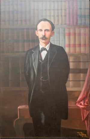 ¾ view of a man with dark hair and a moustache. He wears a black suit and tie and stands in front of a full bookcase.