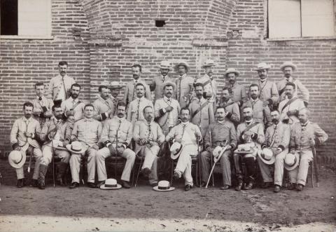 Group of men in light colored clothing posing in front of a stone wall.