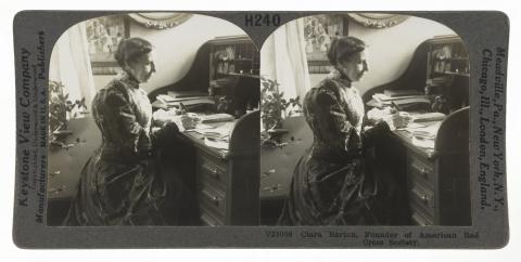  Stereo view of a woman in a long dress seated at a rolltop desk.