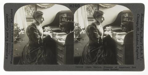 Stereo view of a woman in a long dress seated at a rolltop desk.