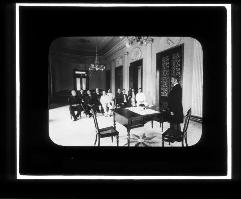 Man standing in a formal room behind a desk with a group of men to his right.