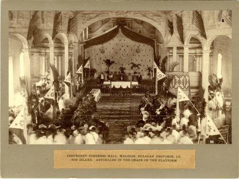 Formal congressional hall with several people in attendance with a man seated on a dais in a chair.